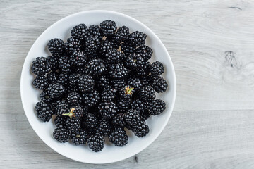 Berries of fresh ripe blackberries in a white plate on a gray wooden background. Healthy summer berries. Healthy vegetarian food. Background with blackberries and free space.