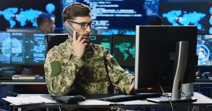 Caucasian male army officer in glasses and uniform talking on telephone and working on computer in analytic secret base in military. Man militarian official in dark office. Digital support. Phone Call