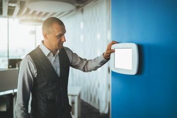 A mature handsome caucasian man entrepreneur using a multifunctional wall-mounted terminal panel with a blank white screen mock-up to adjust illumination level in the office and indoor air temperature