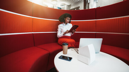 An exquisite black woman entrepreneur sitting on a curved red sofa indoor of a modern office coworking area and using her digital tablet; a paper coffee cup, smartphone, laptop on the table in front