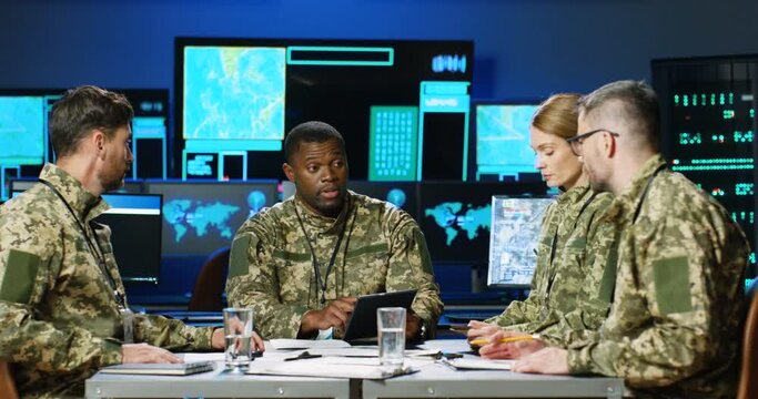 Mixed-races males and female militarian council sitting at table in uniforms, deciding war or defense tactics in dark monitoring room. Multiethnic men and woman officers talking, discussing terrorism.