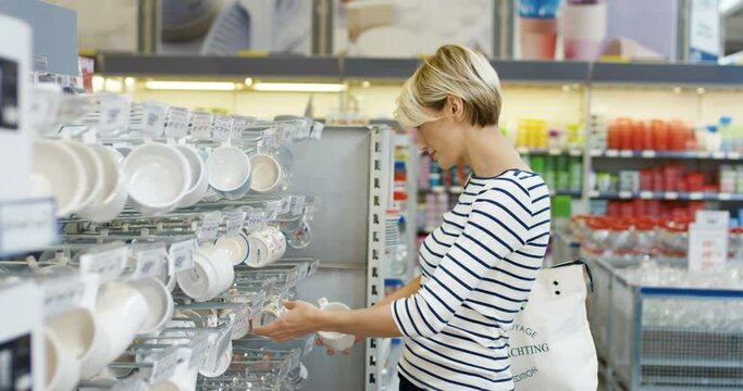 Beautiful Caucasian blonde woman with short hair standing in hypermarket and choosing cups. Indoors. Attractive female client in shop looking for goods for home. Customer buying kitchen items in store
