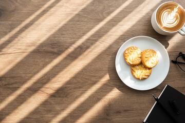 Wood cafe table with dish of almond coffee, cup of coffee, notebook with pen and morning sun light. Top view with copy space, flat lay.