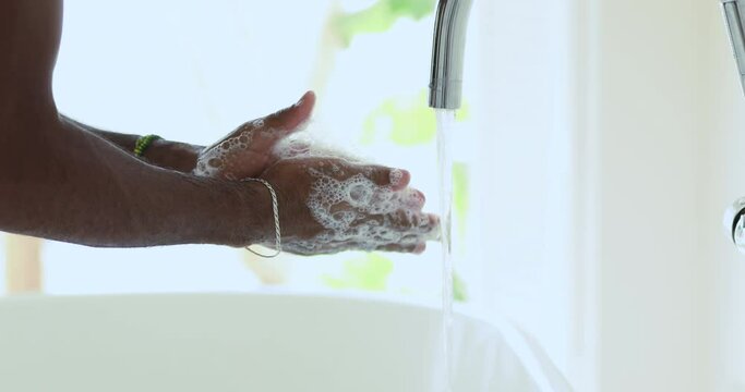 Close up young african american guy rubbing hands with soap over sink with running tap water indoors. Cautious afro man preventing stop spreading coronavirus, sanitizing hands, personal hygiene.
