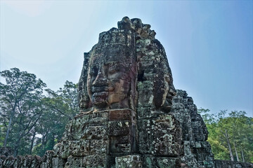 Strange statues at the top of the temple. In the clear sky, huge human faces look in different directions. There are mysterious smiles on their lips. Ancient stones are dilapidated. Cambodia. Angkor.