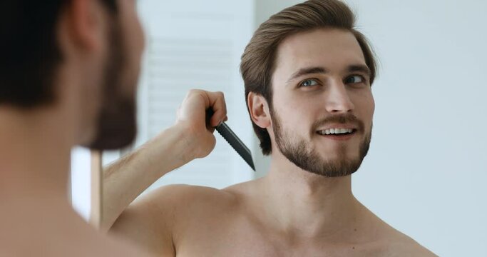 Mirror reflection happy handsome man styling hair with comb, satisfied with stylish haircut. Head shot close up smiling energetic guy getting ready for dating or workday in morning, daily routine.