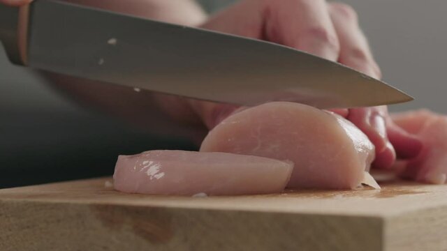 Slow motion man slicing raw chicken fillet on oak cutting board closeup
