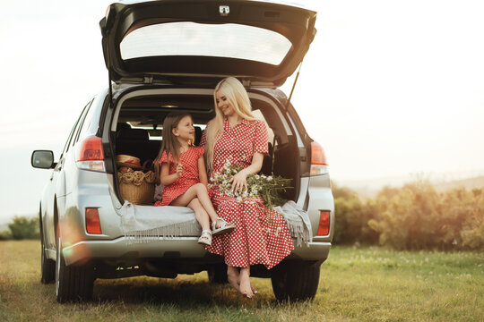 Young Mom with her Little Daughter Dressed Alike in Red Polka Dot Dress, Sitting in a Car Trunk in the Field, Road Trip Picnic Concept