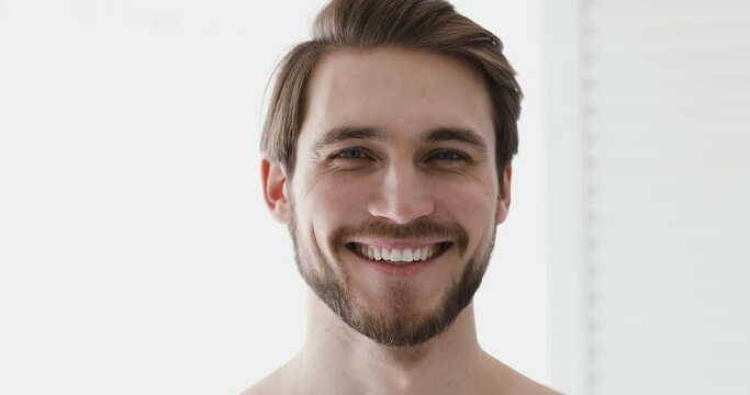 Close up head shot portrait young smiling handsome european appearance man standing in bathroom. Healthy bearded man with healthy white toothy smile looking at camera, feeling energetic in morning.