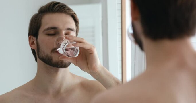 Smiling young healthy bearded guy holding glass, drinking pure fresh water, looking in mirror after showering. Happy handsome man enjoying daily healthcare habit, hydrating thirst in bathroom.
