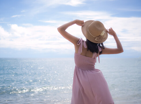 Asian skin tan woman with pink dress and hat she standing relaxing on sand beach with blur image of sea. for travel summer in holidays.