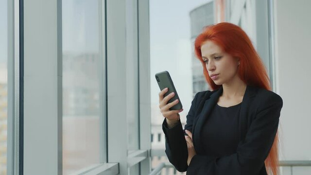 Portrait of a red-haired woman holding a mobile phone in her hand looking gloomily out the window. Angry woman in the office with a cell phone by the window.