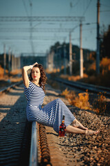 attractive girl in a striped dress and hat sits on the railroad with a bottle of alcohol