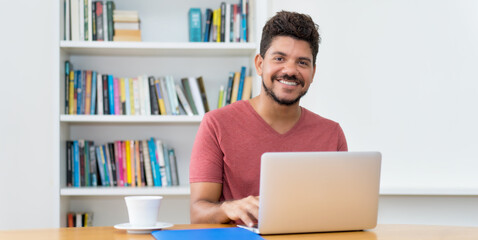 Laughing latin american man with beard working at computer - obrazy, fototapety, plakaty