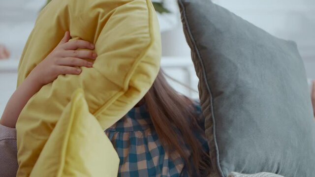 Sweet young beautiful toddler girl with blonde hair hide from her brother covering wigth pillows sitting very quiet. Funny children. Home entertainment. Cozy place.