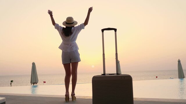 back view of young woman in summer clothes and sun hat, with travel luggage, standing at the pool, enjoying the sunrise. summer. seaside resort. travel concept.