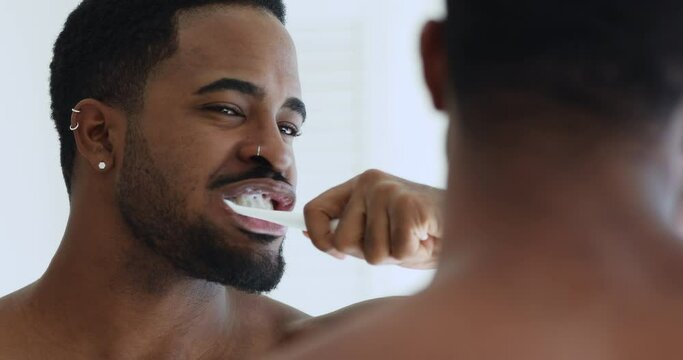 Close up head shot young handsome mixed race man brushing teeth in bathroom. Hipster african ethnicity guy looking in mirror, doing morning oral mouth hygienic daily procedure, toothcare concept.