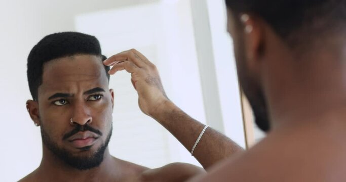 Focused young shirtless african ethnicity man looking in mirror, styling hair with fingers, getting ready in morning. Serious handsome naked mixed race guy doing hairstyle after morning showering.