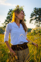 Beautiful romantic woman in a white blouse and long skirt with a wreath of flowers on her head against the background of a rural landscape at sunset. Summer concept. Summer nature, field and trees.