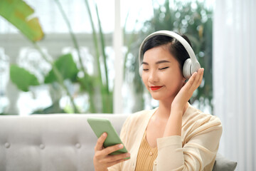 Happy carefree asian girl wearing wireless headphones listening to music with eyes closed sit on sofa,