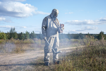 A soldier in a chemical protection suit checks the radioactive territory with a dosimeter against the background of wild vegetation and the sky with clouds. Smoke from harmful fumes is spreading.