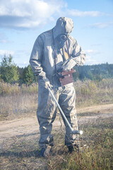 A soldier in a chemical protection suit checks the radioactive territory with a dosimeter against the background of wild vegetation and the sky with clouds. Smoke from harmful fumes is spreading.