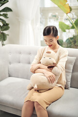 Best friend to hug. Beautiful young asian woman hugging her teddy bear with smile while sitting on couch