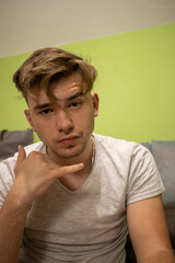 Young male with light brown hair in a white t shirt in a green room holding his hand in a phone position AM