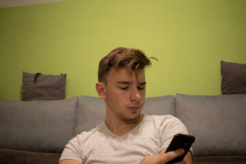 Fototapeta premium Young male with light brown hair in a white t shirt in a green room reading something off his phone AM