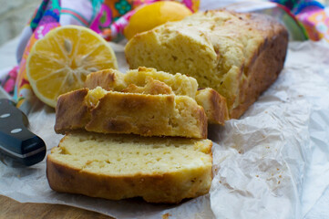 Homemade cake or baking concept. A vanilla or lemon loaf cake freshly baked and sliced on a wooden cutting board. There are lemons and a colourful tea towel in the background. 