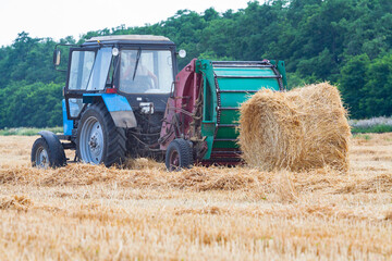Obraz premium tractor makes big straw roll on yellow field at summer day