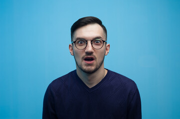 Obraz premium Portrait of an emotional young man in glasses shocked by low prices and discounts on a blue background.