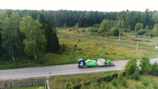 Trucks with oversized cargo travel along the village. Transportation of huge parts. In the background is a dense green forest. Clear summer day.