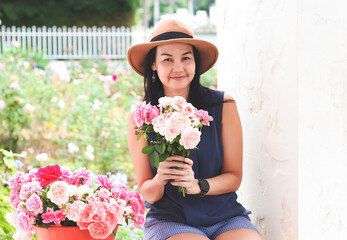 happy Asian woman wearing hat holding roses bouquet,sitting with rose bucket in her rose garden , smiling happily and looking at camera.