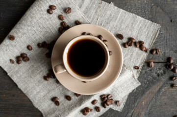 Coffee cup with scattered coffee beans on a wooden table background. Mug of black coffee on a linen textile.
