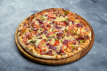 Baked italian pizza with smoked sausages, pickled cucumbers, salami and tomatoes on a wooden tray on a gray background.