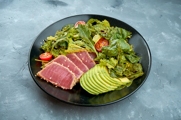 Salad with tuna, avocado and cherry tomatoes in a black plate on a gray background. Close up.