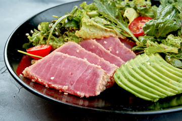 Salad with tuna, avocado and cherry tomatoes in a black plate on a gray background. Close up. Selective focus. Half of plate