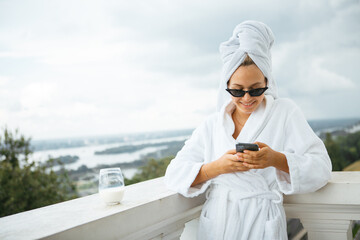 Naklejka premium Smiling cute girl blogger in white robe, towel and sunglasses stands on balcony with phone and cup of milk. Lazy morning