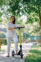 Joyful adorable senior woman using a scooter while riding in the park. Modern woman, a new generation. Healthy cheerful senior retired lady. Concept of Age Inclusivity and ecological transport - 370150345