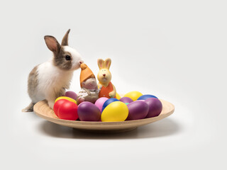 Cute little white and brown rabbit stepping on the wooden plate filled up with colorful eggs, bunny...