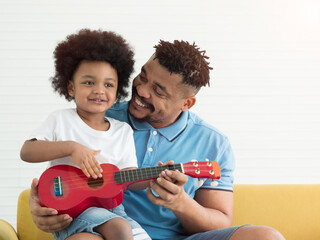 African dad teaching son how to play guitar on the sofa in the living room at home. Father's day,...