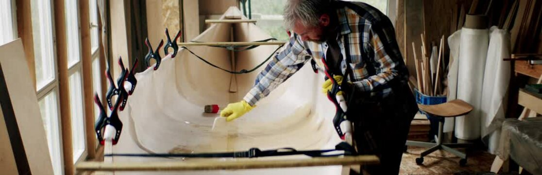 Male building a canoe in his workshop, gluing fiberglass to a plywood frame. Boat making hobby, small business owner