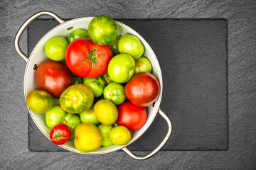 Green and red tomatoes in an enamel sieve on slate slab with copy space