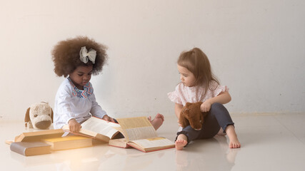 Diverse two cute little toddler girls, African and Caucasian, playing and pretending to read thick books in a white room.