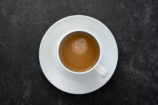 A flat lay top down photo of a white saucer and cup filled with coffee placed on dark stone table