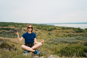 Naklejka na ściany i meble A Caucasian woman with a drone in her hand, sitting on a green rocky hill with sky in background