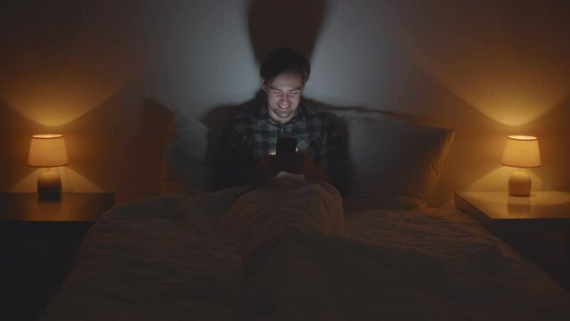 Smiling young man lying on bed at home at night using mobile phone