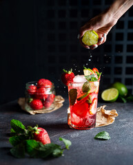 Strawberry lime lemonade in glass decorated with mint. Drops is falling from the squeezed lime in the woman's hand