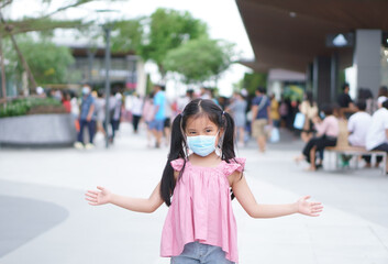 Fototapeta premium Asian child or kid girl smile wear paper face mask and open arm to present on lot of people for social distancing from person enjoy shopping at department store or market mall on holiday in covid era
